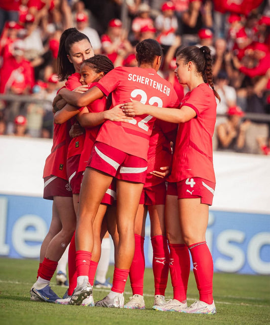 Group of female soccer players in red uniforms celebrating on a field with a crowd in the background.